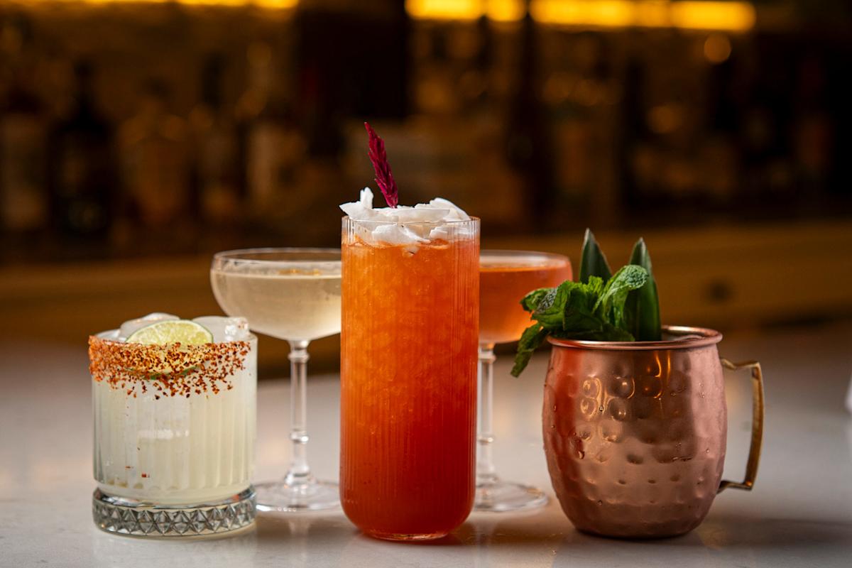 A lineup of four assorted cocktails in different glasses sits on a bar, including a salted-rim drink with lime, a coupe glass with a pale cocktail, a tall orange drink with ice and garnish, and a copper mug with mint leaves.