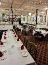 A cozy restaurant dining room with long tables set for a meal, featuring white tablecloths, red napkins, wooden chairs, string lights, and walls decorated with framed art and various hanging plants.