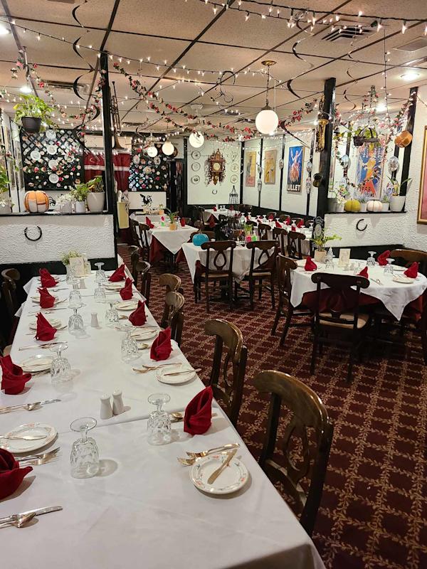A cozy restaurant dining room with long tables set for a meal, featuring white tablecloths, red napkins, wooden chairs, string lights, and walls decorated with framed art and various hanging plants.