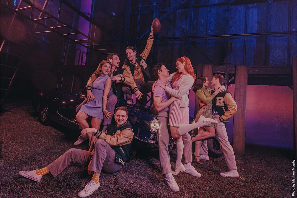 Seven young adults in vintage clothes pose cheerfully around a classic car under purple lighting. Some wear letterman jackets; two people embrace, and another holds a football up high. The scene feels playful and theatrical.