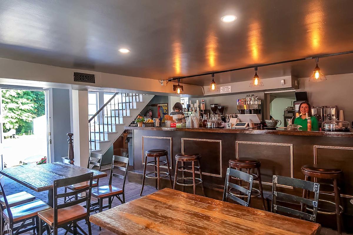 A cozy café interior with wooden tables and chairs, a counter with bar stools, coffee machines, and a smiling barista standing behind the counter under warm lighting. A staircase and open door are visible in the background.