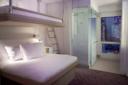 A modern hotel room with a double bed, an overhead bunk, a glass-enclosed shower, floor-to-ceiling windows with city views, white walls, and minimalistic decor.