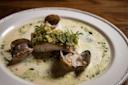 A plated dish featuring a fillet of white fish topped with a green garnish, surrounded by clams in a creamy, foamy sauce, served on a white plate with a decorative edge on a wooden table.