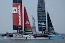 Two racing sailboats with "ROCKWOOL" and "BLACK FOILS" sails compete on the water near the Statue of Liberty, with the iconic monument visible in the background on a clear day.