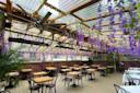 A charming outdoor patio with wooden tables and black chairs under a transparent roof. Purple wisteria flowers hang from the ceiling. The flooring is wooden, and a small plant is visible. A person stands in the background.