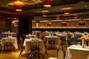 Elegant restaurant interior with neatly set tables covered in white cloths, classic black chairs, warm lighting from red lampshades, wooden floors, and decorative wallpaper with framed art on the walls.