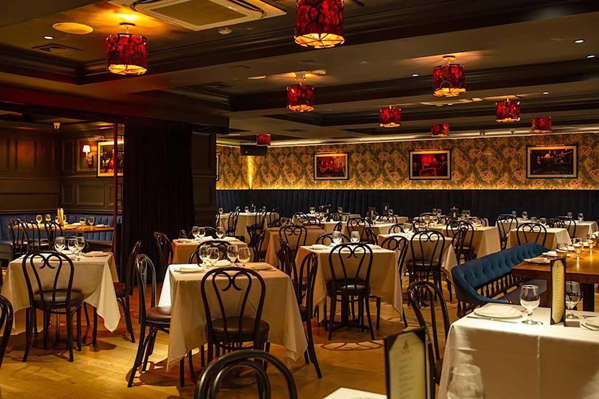 Elegant restaurant interior with neatly set tables covered in white cloths, classic black chairs, warm lighting from red lampshades, wooden floors, and decorative wallpaper with framed art on the walls.