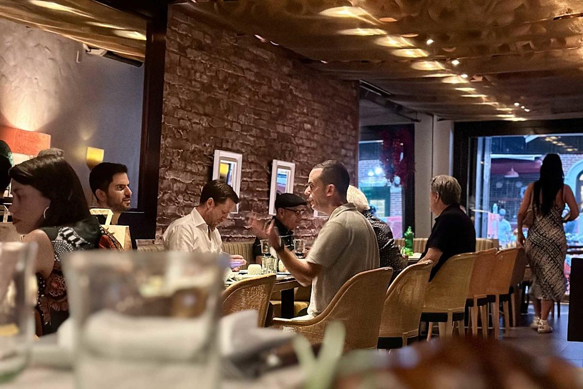 People are dining and conversing at a cozy restaurant with warm lighting, brick walls, and framed art. Some patrons are eating, while others chat. A woman in a patterned dress walks toward the entrance.