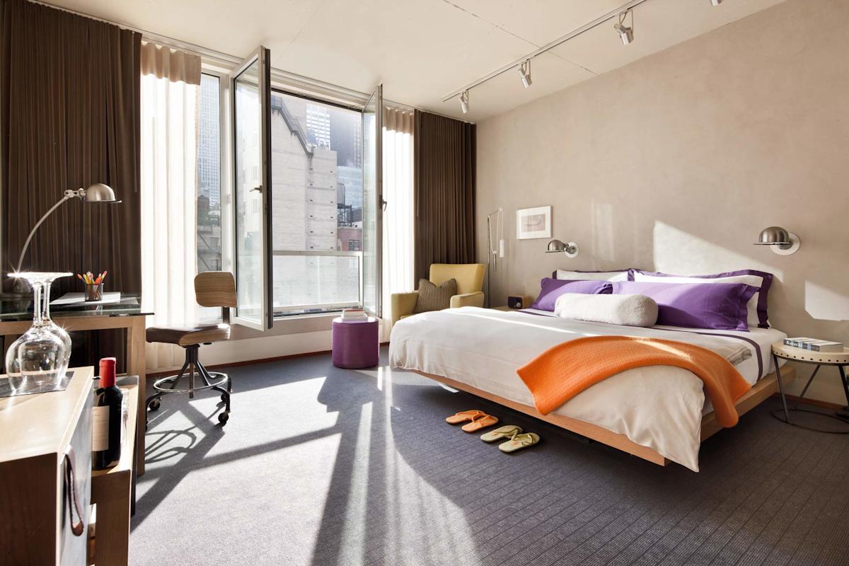 Modern bedroom with large windows, open to city view. Bed with white linens, purple pillows, orange throw. Desk, chair, wine, glass, and slippers on the floor. Cozy reading nook with yellow chair and art on the wall.