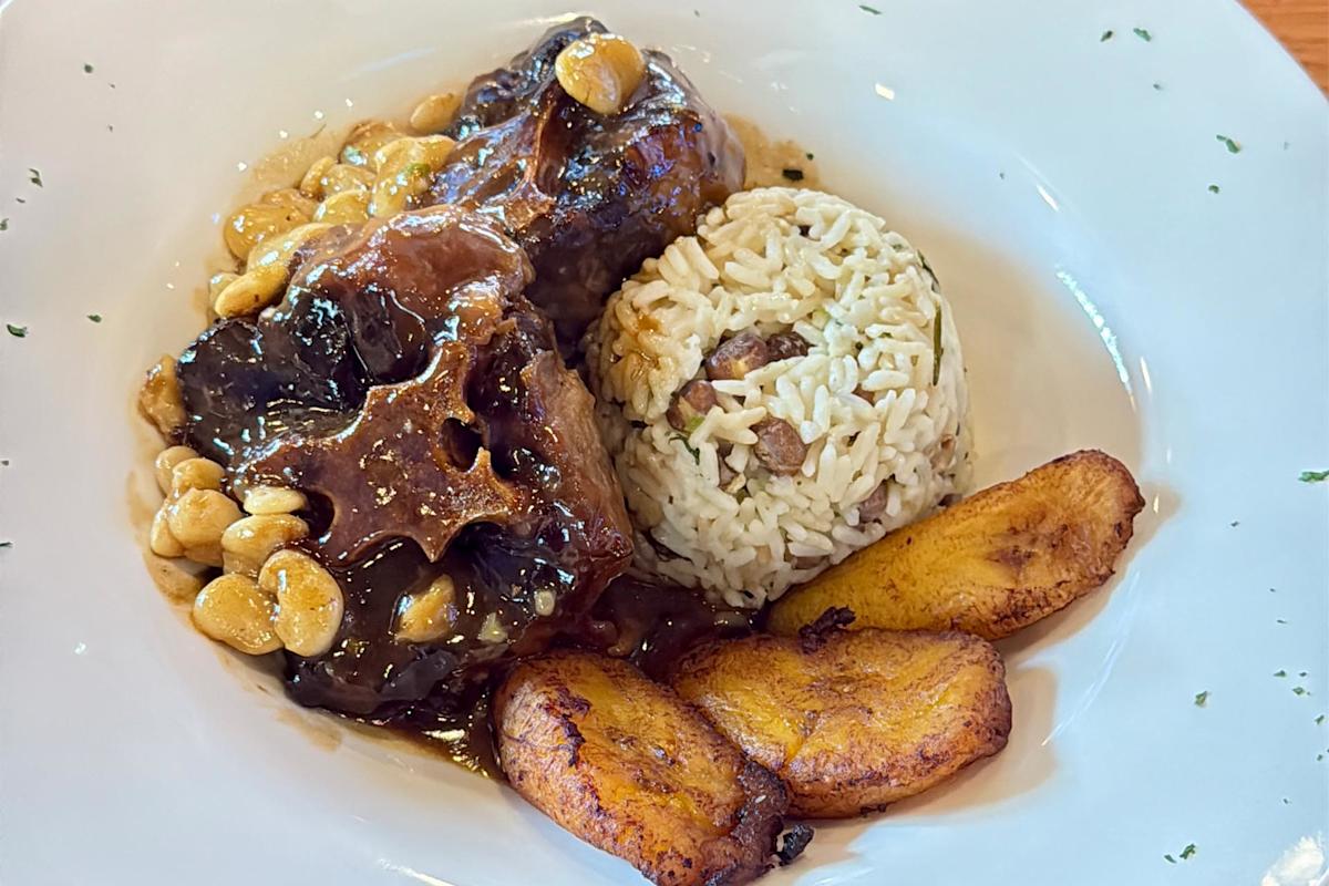 A plate with braised oxtail topped with beans, a mound of rice and peas, and two pieces of fried plantain, served on a white dish.
