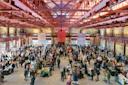 Spacious indoor market with high ceilings and red beams. A large crowd moves between vendor booths filled with various goods, under natural light streaming through windows. Walls are adorned with art and murals.