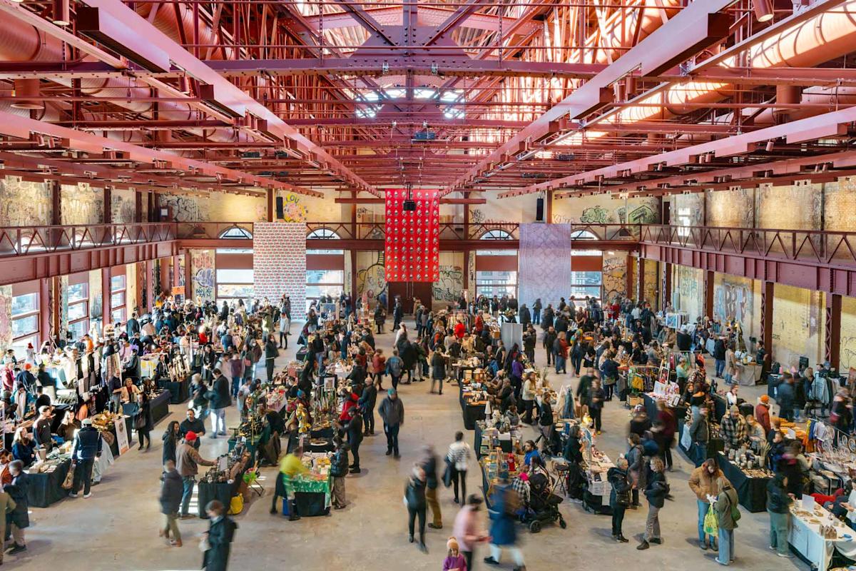 Spacious indoor market with high ceilings and red beams. A large crowd moves between vendor booths filled with various goods, under natural light streaming through windows. Walls are adorned with art and murals.