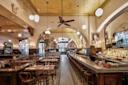 Interior of a vintage-style restaurant with white tiled walls, arched mirrors, and globe pendant lights. Wooden tables and chairs line the floor along with a long bar. A large archway leads to the back, with a chandelier and greenery visible.