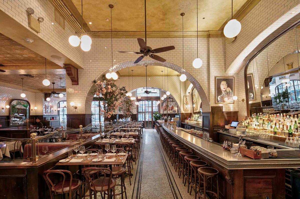 Interior of a vintage-style restaurant with white tiled walls, arched mirrors, and globe pendant lights. Wooden tables and chairs line the floor along with a long bar. A large archway leads to the back, with a chandelier and greenery visible.