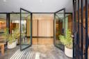 Modern building entrance with large glass double doors open, wood paneling on the back wall, potted green plants on either side, and a concrete floor. The space looks clean and inviting.