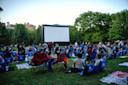 A crowd of people sits on lawn chairs and blankets in a grassy park, facing a large outdoor movie screen, surrounded by trees under a clear sky.
