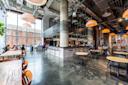 A modern cafe with high ceilings, large windows, hanging lights, and industrial decor. People sit at wooden and metal tables, chatting and working. The bar area is central, with exposed pipes overhead.
