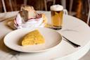 A slice of Spanish tortilla sits on a white plate, with a glass of beer and a small basket of bread wrapped in a striped cloth on a marble table. A fork is placed beside the plate.