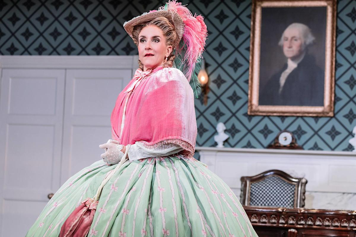 A person in an elaborate, pastel-colored 18th-century costume and hat stands in a vintage room with patterned wallpaper, a wooden chair, and a portrait of George Washington on the wall.