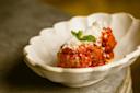 A decorative white bowl holds two meatballs covered in tomato sauce and sprinkled with grated cheese. A small mint leaf garnishes the dish, set on a gray surface.