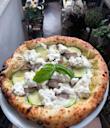A freshly baked pizza topped with zucchini slices, meat, dollops of ricotta cheese, and a fresh basil leaf, held up in a cozy, plant-filled restaurant with tables and chairs in the background.