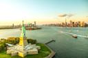 Aerial view of the Statue of Liberty on Liberty Island with New York City skyline in the background. The sunset casts a warm glow over the city and the water. Several boats are visible in the harbor.