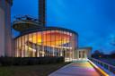 A modern building with large curved glass walls is illuminated with colorful lights at dusk. A tall structure with a circular platform is visible in the background. The sky is deep blue, adding contrast to the scene.
