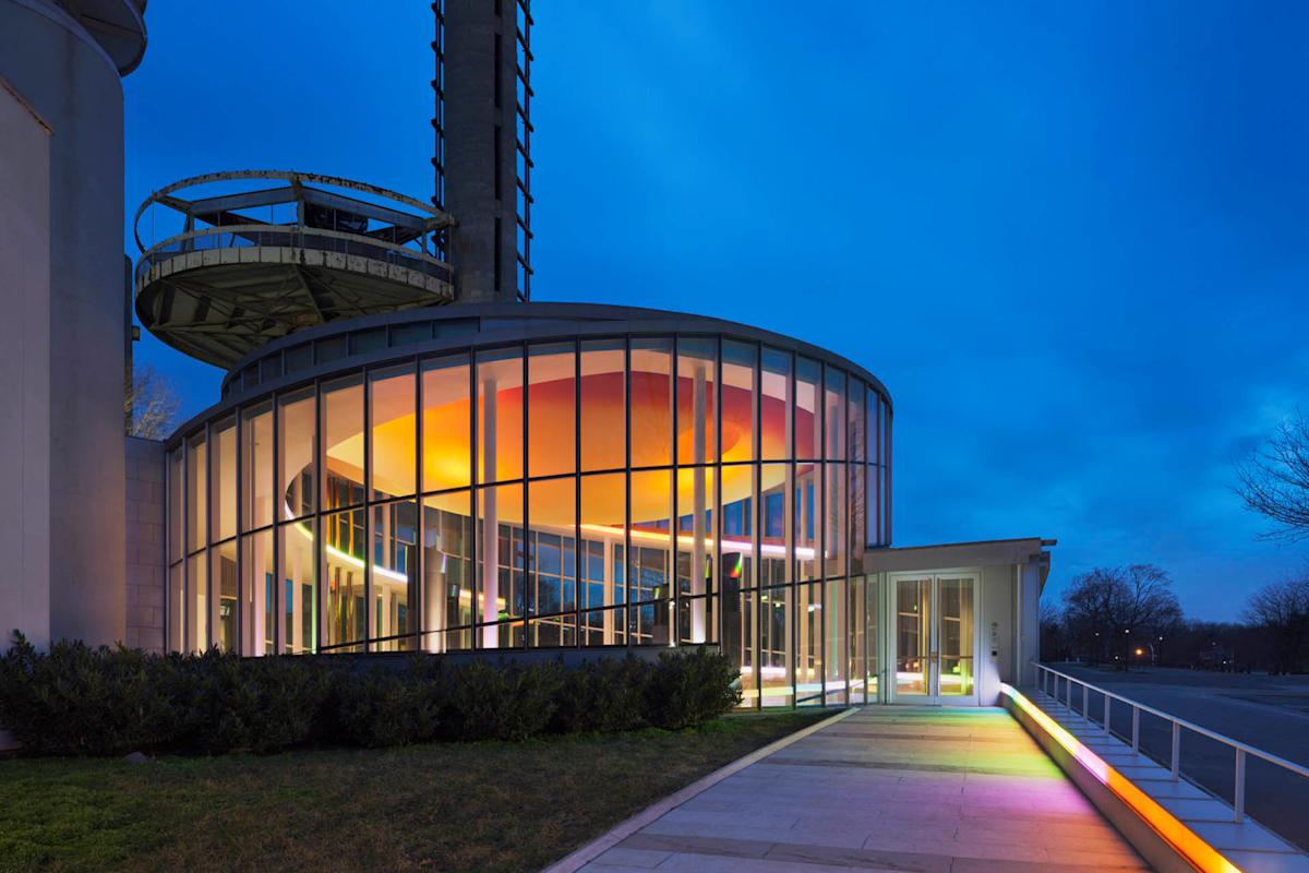 A modern building with large curved glass walls is illuminated with colorful lights at dusk. A tall structure with a circular platform is visible in the background. The sky is deep blue, adding contrast to the scene.