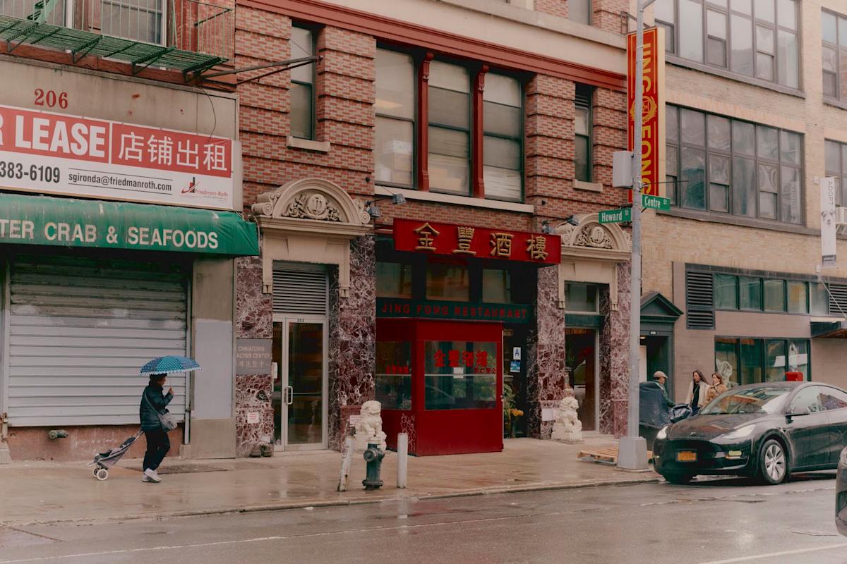 A person with an umbrella walks past closed and open shops on a rainy city street, including a restaurant with Chinese signage and a "FOR LEASE" sign in English and Chinese. A parked car and another person are also visible.