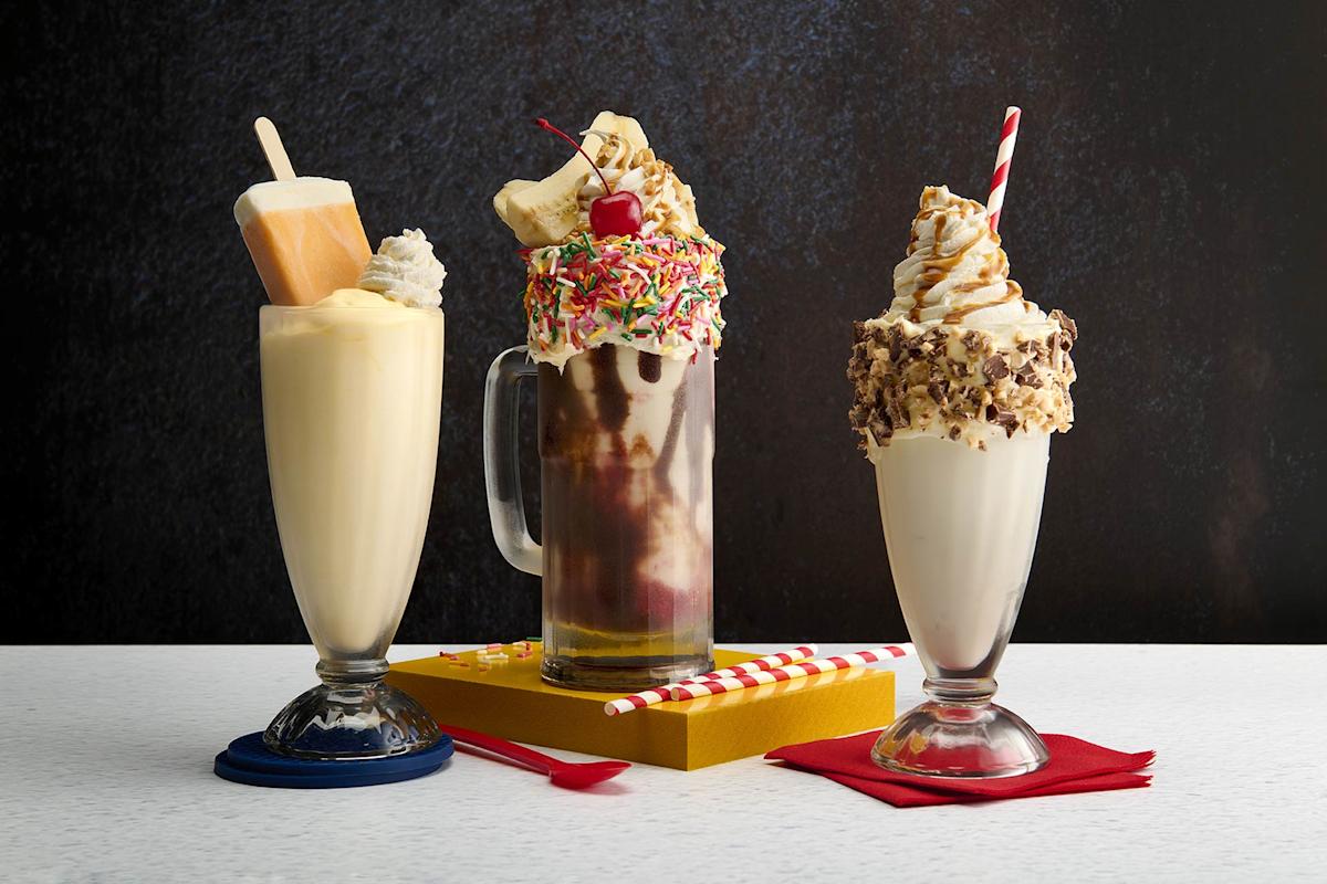 Three over-the-top milkshakes: one with a popsicle and whipped cream, one with chocolate, bananas, rainbow sprinkles, and a cherry, and one with whipped cream, caramel drizzle, and chocolate pieces, against a dark background.