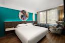 A modern hotel room with a large bed, teal accent wall, abstract circular artwork above the headboard, two bedside tables, a patterned armchair, and a large window with sheer curtains.