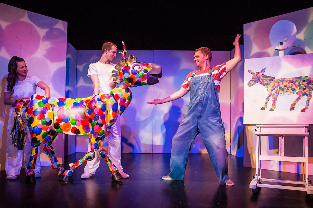 Three people on stage with a colorful spotted donkey figure; one person wears overalls, another holds the donkey, and a third stands nearby. A painting of the spotted donkey is on an easel.