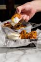 A hand dips a saucy buffalo chicken wing into a cup of creamy ranch dressing, with fries and more wings in a basket lined with newspaper-style paper on a marble table.