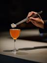 A hand uses metal tweezers to place an edible flower garnish on an orange cocktail in a stemmed glass, set against a dark background.