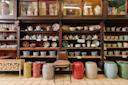 Shelves filled with colorful ceramic bowls, plates, and teapots line a wooden wall. Patterned stools and chairs are arranged in front, and various decorative items are displayed above the shelves.