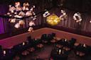 A dimly lit, elegant restaurant interior with plush, curved seating along the walls. Tables are set with candles and glassware. Beautiful floral-shaped chandeliers hang from the ceiling, casting warm, ambient light.
