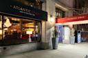 Exterior of Atlantic Grill at night, with people visible inside through large windows, a chandelier hanging, and potted plants by the entrance. Sign above reads "ATLANTIC GRILL Born in New York 1998.