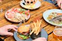 A wooden table set with a burger and fries, a glass of beer, a plate of shrimp with lemon wedges, a platter of bread and cured meats, and a plate with grilled fish and vegetables. Hands reach for the food.