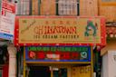 A storefront sign for the Original Chinatown Ice Cream Factory with a cartoon dragon holding ice cream, red and yellow lettering, and neon signs in English and Chinese. The shop is located at 65 Bayard Street.