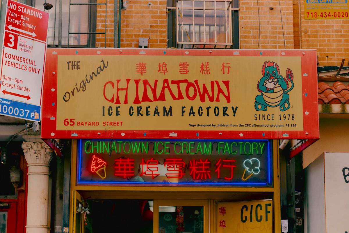 A storefront sign for the Original Chinatown Ice Cream Factory with a cartoon dragon holding ice cream, red and yellow lettering, and neon signs in English and Chinese. The shop is located at 65 Bayard Street.