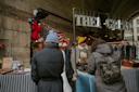Two people in warm coats browse a stall at an outdoor market. The vendor smiles, wearing a yellow hat. The stall, decorated with festive lights and an American flag, displays various items. A sign above reads "THEA GRANT.