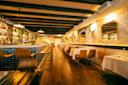 A warmly lit, modern restaurant with wooden floors, tan chairs, white tablecloths, and a bar with high stools. Bottles line the shelves, and decorative plants hang on the walls. The space appears inviting and cozy.