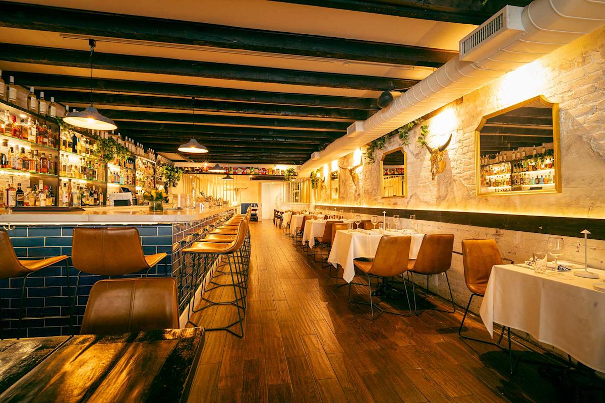 A warmly lit, modern restaurant with wooden floors, tan chairs, white tablecloths, and a bar with high stools. Bottles line the shelves, and decorative plants hang on the walls. The space appears inviting and cozy.