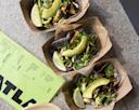 Three tacos in brown paper trays, each filled with greens, fried filling, avocado slices, and a lime wedge, are arranged on a speckled countertop next to a green menu.