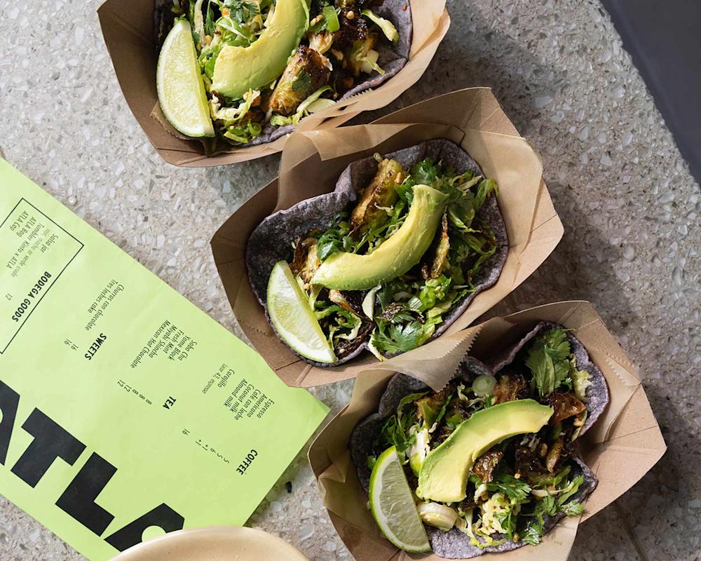 Three tacos in brown paper trays, each filled with greens, fried filling, avocado slices, and a lime wedge, are arranged on a speckled countertop next to a green menu.