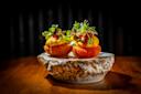 Two artfully plated bite-sized appetizers topped with microgreens, creamy sauce, and garnished with red accents sit on a textured ceramic dish against a dark background, lit dramatically.