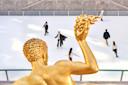 View of an ice rink from above a golden statue. Several people are skating, wearing winter clothing. The scene is set against a gray stone wall backdrop, capturing a bright and lively atmosphere.