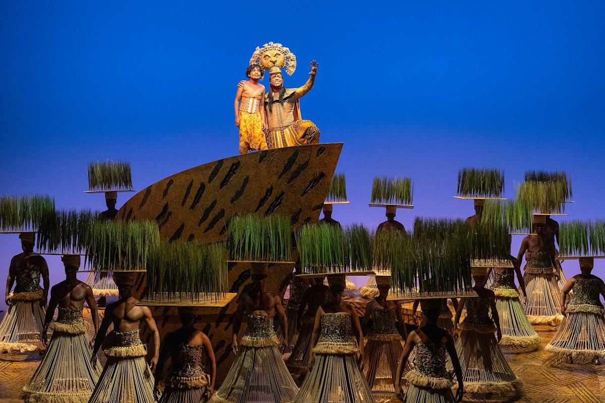 Performers in colorful costumes stand on a decorated platform, resembling a lion and cub, with other dancers below, wearing headpieces topped with grass, against a vivid blue background in a stage production.