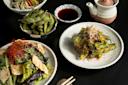 A selection of Japanese dishes on a black table, including a salad with greens and crispy toppings, a bowl of edamame, grilled shishito peppers with bonito flakes, dipping sauce, and a teapot.