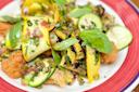 Colorful salad with grilled yellow squash, zucchini ribbons, fresh basil leaves, chopped herbs, diced red onions, and crispy bread pieces on a red and white plate.